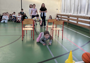 Dziecko czołga się pod krzesłami podczas toru przeszkód na sali gimnastycznej, a grupa uczniów świetlicy obserwuje i kibicuje.