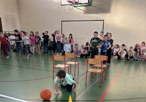 Dziecko czołga się po podłodze sali gimnastycznej, omijając pachołek i piłkę podczas zawodów sportowych, w tle grupa uczniów kibicuje.