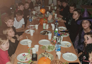 Uczniowie klasy 4a siedzą przy długim stole podczas halloweenowej zabawy w sali lekcyjnej; na stole znajdują się przekąski, napoje i dynie, a dzieci mają różne przebrania i makijaże.