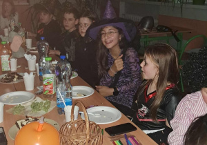 Uczniowie w halloweenowych przebraniach siedzą przy stole podczas klasowej zabawy; na stole znajdują się talerzyki, słodycze, napoje i dekoracje, a jedna z dziewczynek w czarodziejskim kapeluszu uśmiecha się do aparatu.