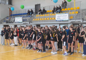 Grupa uczniów w czarnych koszulkach ustawiona na parkiecie hali sportowej podczas charytatywnego meczu wolontariuszy szkół pabianickich, w tle trybuny i banery sponsorów.