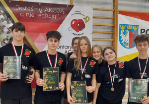 Uczniowie SP 13 w Pabianicach stojący z medalami i dyplomami na tle banerów charytatywnego turnieju w hali sportowej.