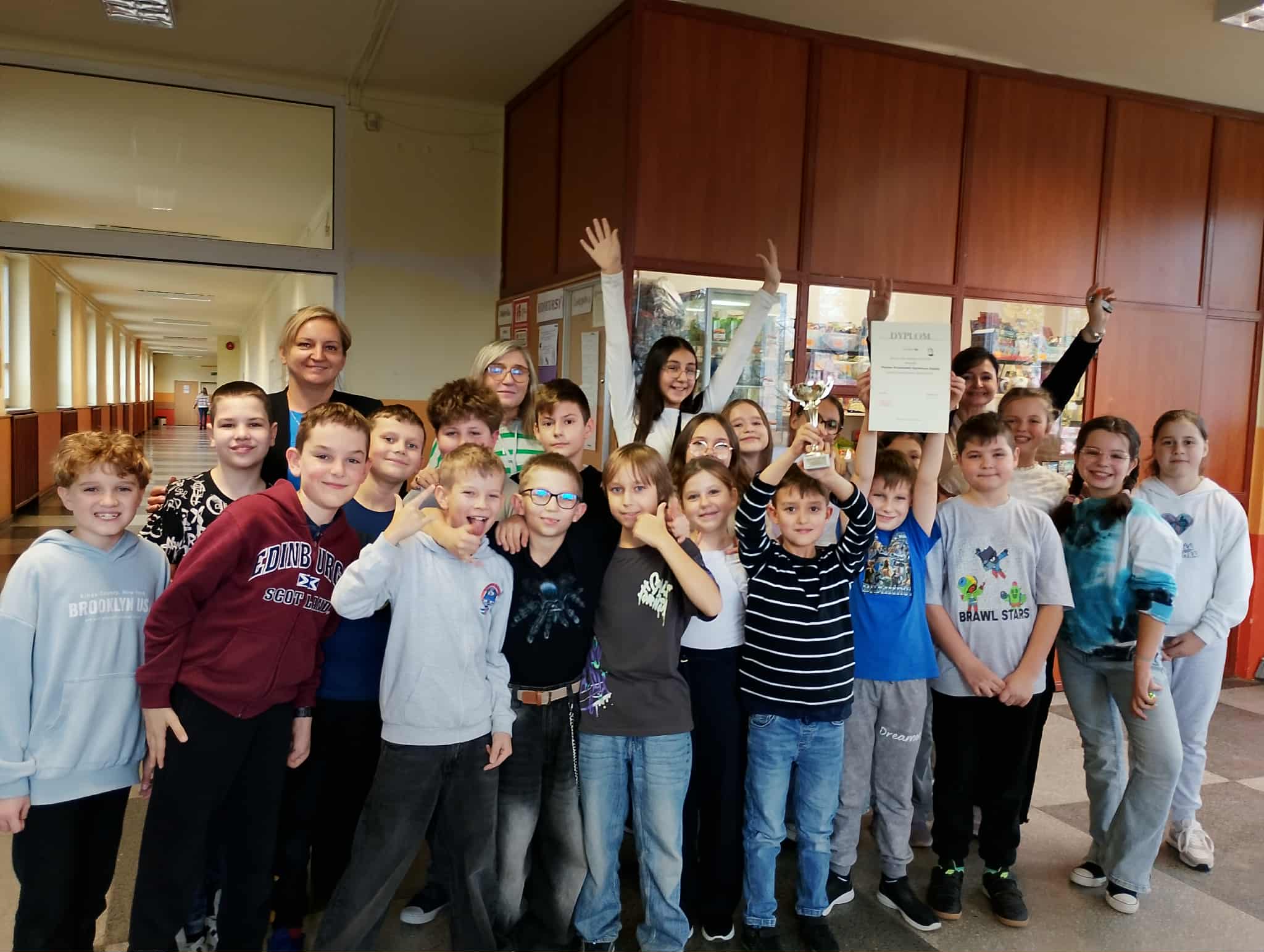 Uczniowie klasy 4a z SP 13 w Pabianicach stoją na korytarzu, trzymają puchar i dyplom za zwycięstwo w Stefankach, uśmiechając się i pozując razem z Dyrektorką Szkoły.