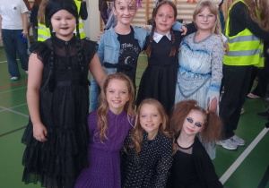 Siedem uczennic klasy 4 w halloweenowych przebraniach pozuje na sali gimnastycznej; mają mroczne i fantazyjne stroje oraz makijaże, stojąc i siedząc w dwóch rzędach.