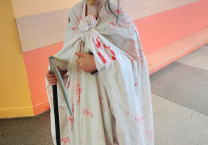 Uczeń klasy 4 w halloweenowym kostiumie z dużą straszną maską i białym prześcieradłem z czerwonymi plamami, trzymająca czarną laskę na szkolnym korytarzu.