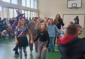 Grupa uczniów tańczy na sali gimnastycznej podczas dyskoteki halloweenowej; niektóre dzieci mają przebrania, a w tle siedzą inni uczniowie.