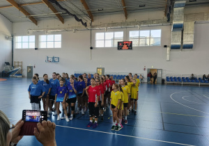 Drużyny dziewcząt w różnych kolorach strojów ustawione w kolumnach na hali sportowej; z prawej strony idą zawodniczki SP 13 w Pabianicach w żółtych koszulkach.
