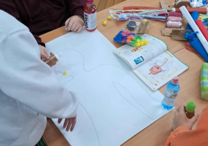 Uczniowie przy ławce rysują sylwetkę człowieka na arkuszu papieru, przygotowując się do wykonania modelu układu pokarmowego z różnorodnych materiałów.