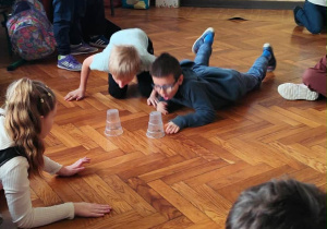 Uczniowie klasy 2a biorą udział w wyścigu na brzuchach po parkiecie, ścigając się do plastikowych kubeczków podczas andrzejek.