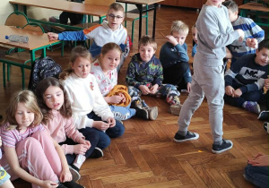 Grupa dzieci z klasy 2a siedzi na parkiecie w kole, przed nimi znajduje się metalowa miska z wodą do andrzejkowych zabaw.