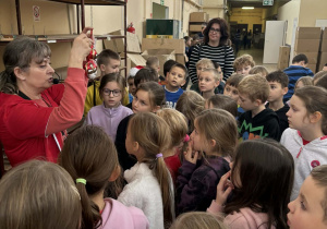 Dzieci z klas 2a i 2b słuchają pracownicy fabryki, która prezentuje im gotową świąteczną bombkę w czasie zwiedzania zakładu.