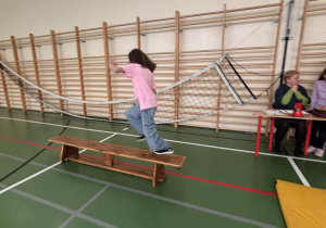 Dziewczynka idzie po ławce gimnastycznej przy drabinkach, a nauczycielki przy stoliku obserwują jej start w olimpiadzie świetlicowej.