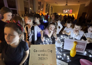 Dzieci w świetlicy uczestniczą w andrzejkowych wróżbach przy stoliku z lampkami, na pierwszym planie karta „Co czeka Cię w roku 2026?” z kolorowo zaznaczonymi liczbami.