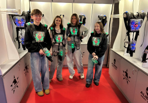 Uczniowie klasy 7a w kamizelkach z sensorami i z pistoletami laserowymi pozują przed wejściem na arenę Laser Games podczas klasowych mikołajek.