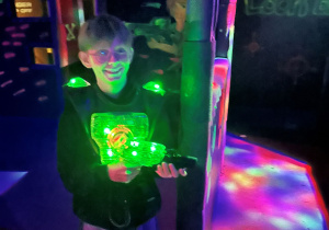 Uczeń klasy 7a na arenie Laser Games trzyma podświetlony na zielono pistolet laserowy, przygotowany do oddania kolejnego strzału.