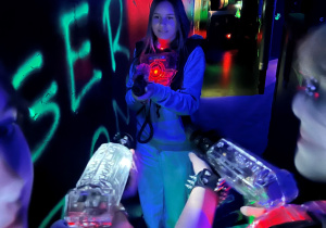 Uczennica klasy 7a w labiryncie Laser Games celuje z pistoletu laserowego, otoczona przez graczy z wycelowaną w nią bronią w kolorowych światłach areny.