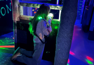 Uczennica 7a klęczy za osłoną na arenie Laser Games i mierzy z pistoletu laserowego w półmroku z kolorowymi światłami.