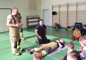 Na sali gimnastycznej odbywa się pokaz udzielania pierwszej pomocy. Dwóch strażaków, jeden stojący, drugi klęczący, prowadzą instruktaż dla grupy dzieci siedzących na podłodze. Trzeci strażak leży na macie, symulując osobę poszkodowaną. W tle widoczne są drabinki gimnastyczne, sprzęt ratowniczy i kurtki. Dzieci uważnie obserwują demonstrację.