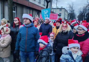 Tłum dzieci i dorosłych w czerwonych czapkach Mikołaja maszeruje ulicą podczas Pabianickiej Parady Świątecznej, w środku widoczny szyld Szkoła Podstawowa nr 13.