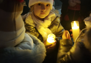 Uczennica SP 13 w świątecznej czapce trzyma zapaloną świecę, stojąc w grupie dzieci podczas Pabianickiego Jarmarku Świątecznego.