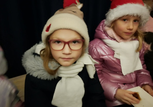 Dwie dziewczynki w zimowych kurtkach, białych szalikach i świątecznych czapkach siedzą obok siebie, w tle widać innych uczestników jarmarku.