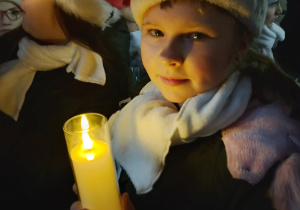Dziewczynka z SP 13 w czapce Mikołaja trzyma zapaloną świecę podczas wieczornego fragmentu jarmarku.