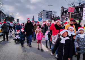 Grupa dzieci i dorosłych w zimowych kurtkach i czapkach Mikołaja idzie środkiem ulicy w świątecznej paradzie, część uczestników macha do obiektywu.