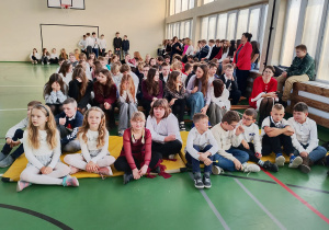 Uczniowie klas 4–8 siedzą w sali gimnastycznej podczas szkolnej uroczystości.