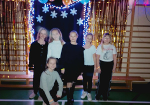Sześć uczennic pozuje na tle dekoracji z balonami, śnieżynkami i światełkami podczas szkolnej dyskoteki karnawałowej. Dziewczynki ubrane w dyskotekowe stroje, uśmiechają się do zdjęcia w udekorowanej sali gimnastycznej.