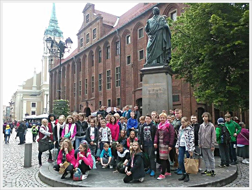Wycieczka uczni&oacute; klas młodszych do Torunia. Grupa dzieci i opiekun&oacute;w pozuje do zdjęcia grupowego przed pomnikiem Mikołaja Kopernika w Toruniu. Pomnik znajduje się na wysokim cokole z inskrypcją łacińską. W tle widoczna jest ceglana fasada Ratusza Staromiejskiego z arkadowymi oknami oraz wieża kościoła św. Ducha. Na placu widać brukowaną nawierzchnię, drzewa oraz tłumy turyst&oacute;w. Zdjęcie zrobione podczas wycieczki szkolnej.
