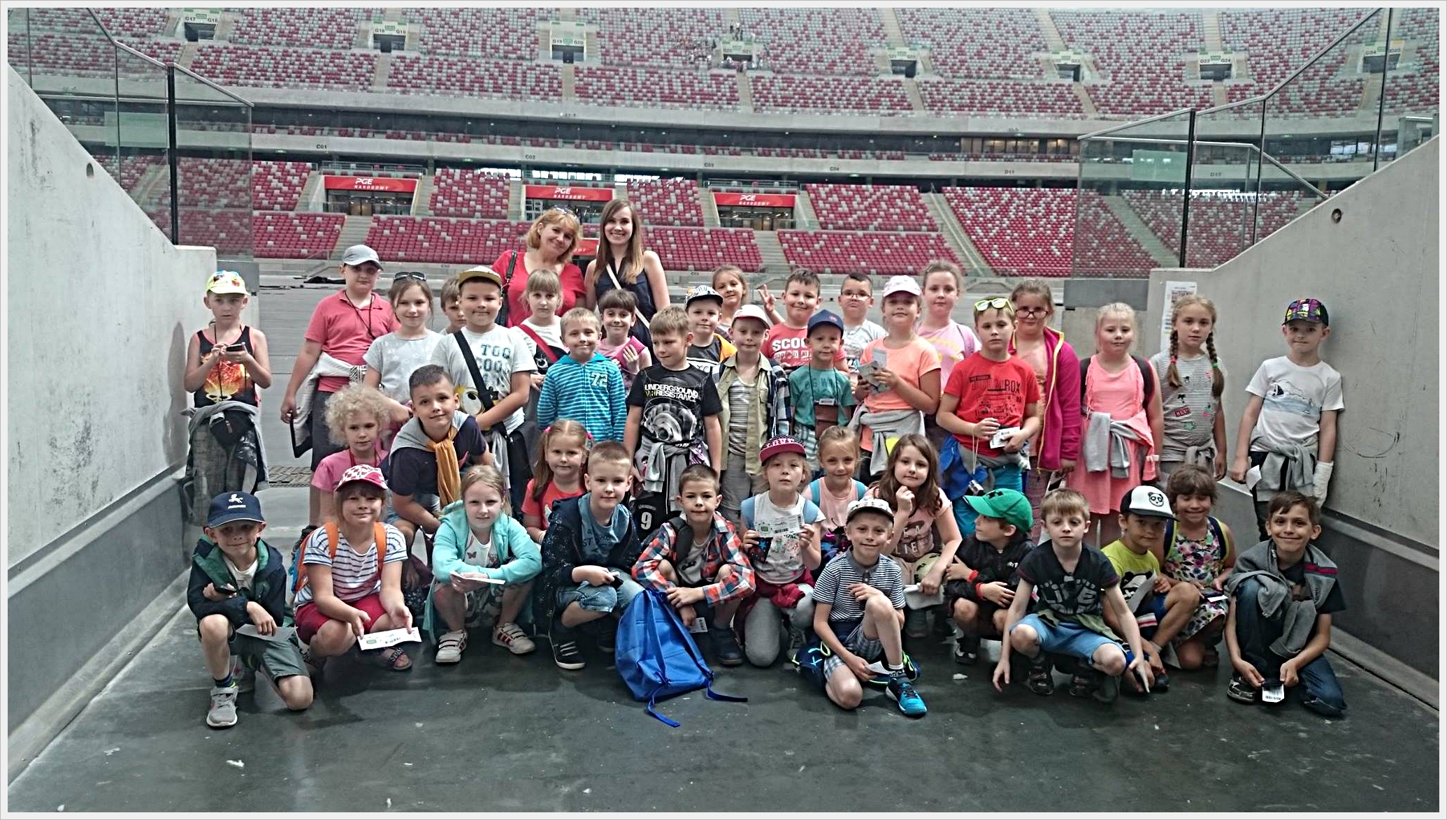 Wycieczka klas trzecich do Warszawy Duża grupa dzieci pozuje do wsp&oacute;lnego zdjęcia na tle stadionu z czerwono-białymi miejscami na trybunach. Dzieci stoją i kucają między betonowymi ścianami w tunelu prowadzącym na murawę. W tle widać ogrom trybun, co sugeruje, że zdjęcie wykonano na dużym stadionie sportowym, prawdopodobnie podczas wycieczki szkolnej. Dzieci są ubrane letnio, wiele z nich ma czapki z daszkiem, plecaki i bilety w rękach. Towarzyszą im dwie uśmiechnięte opiekunki.