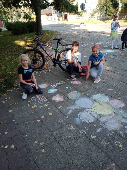 Niecodzienna lekcja. Trzy dziewczynki siedzą na chodniku i uśmiechają się do zdjęcia, otoczone kolorowymi rysunkami wykonanymi kredą. Na chodniku widać m.in. rysunek dużego kwiatka i r&oacute;żne kształty w intensywnych kolorach. W tle stoi rower, a po prawej stronie widać inne dzieci oraz drzewa rzucające cień na chodnik.