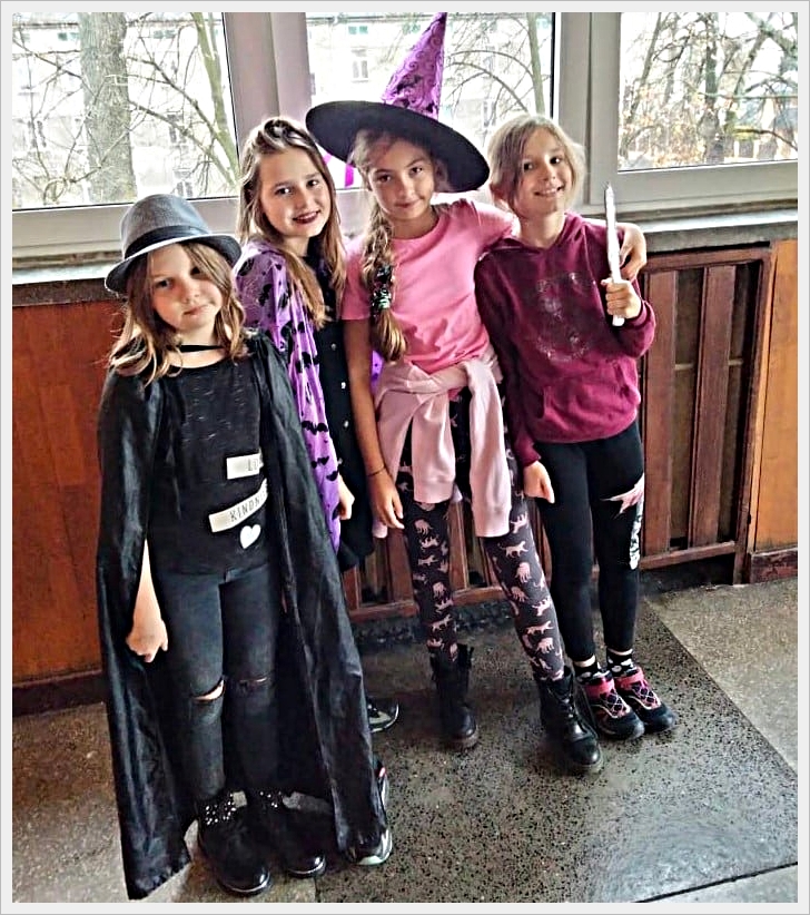 Ostatki w naszej szkole. Cztery dziewczynki stoją obok siebie w szkolnym korytarzu, przebrane w kolorowe stroje, prawdopodobnie z okazji Halloween lub balu przebierańc&oacute;w. Pierwsza z lewej ma na sobie czarne ubranie z peleryną, kapelusz i napisy na koszulce. Druga dziewczynka nosi fioletową pelerynę w czarne nietoperze. Trzecia jest ubrana w r&oacute;żową bluzkę, legginsy z r&oacute;żowymi kotami i ma na głowie duży kapelusz czarownicy. Czwarta dziewczynka ubrana jest w bordową bluzę i trzyma w ręku r&oacute;żdżkę. Wszystkie stoją w uśmiechniętej pozie na tle okien z widokiem na drzewa i budynki za nimi.