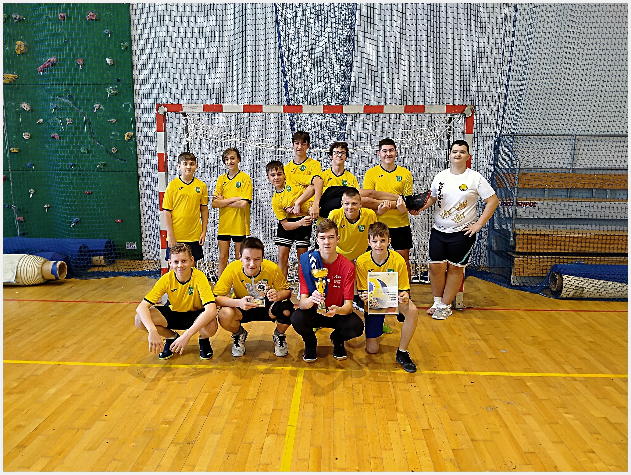 Zawody sporotwe - piłka ręczna. Grupa chłopc&oacute;w w ż&oacute;łtych koszulkach sportowych pozuje w sali gimnastycznej na tle bramki do piłki ręcznej. Trzech z nich klęczy z przodu, trzymając puchar i dyplom. W tle widać ściankę wspinaczkową, siatki zabezpieczające i zrolowane maty gimnastyczne. Wszyscy są uśmiechnięci i wyglądają na zadowolonych z osiągnięcia sportowego.