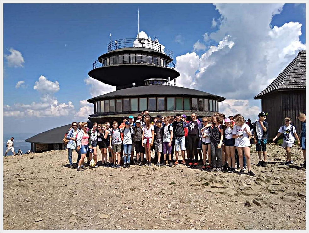 Wycieczka klas &oacute;smych - zdobycie Śniezki. Grupa młodzieży pozuje do zdjęcia na szczycie g&oacute;ry przed charakterystycznym, futurystycznym budynkiem o okrągłych kondygnacjach &ndash; Obserwatorium Meteorologicznym na Śnieżce. Uczniowie stoją na kamienistym podłożu, ubrani w letnie stroje turystyczne, niekt&oacute;rzy mają plecaki i czapki z daszkiem. W tle widać niebo z chmurami oraz fragment drewnianego schroniska. Panuje słoneczna pogoda.