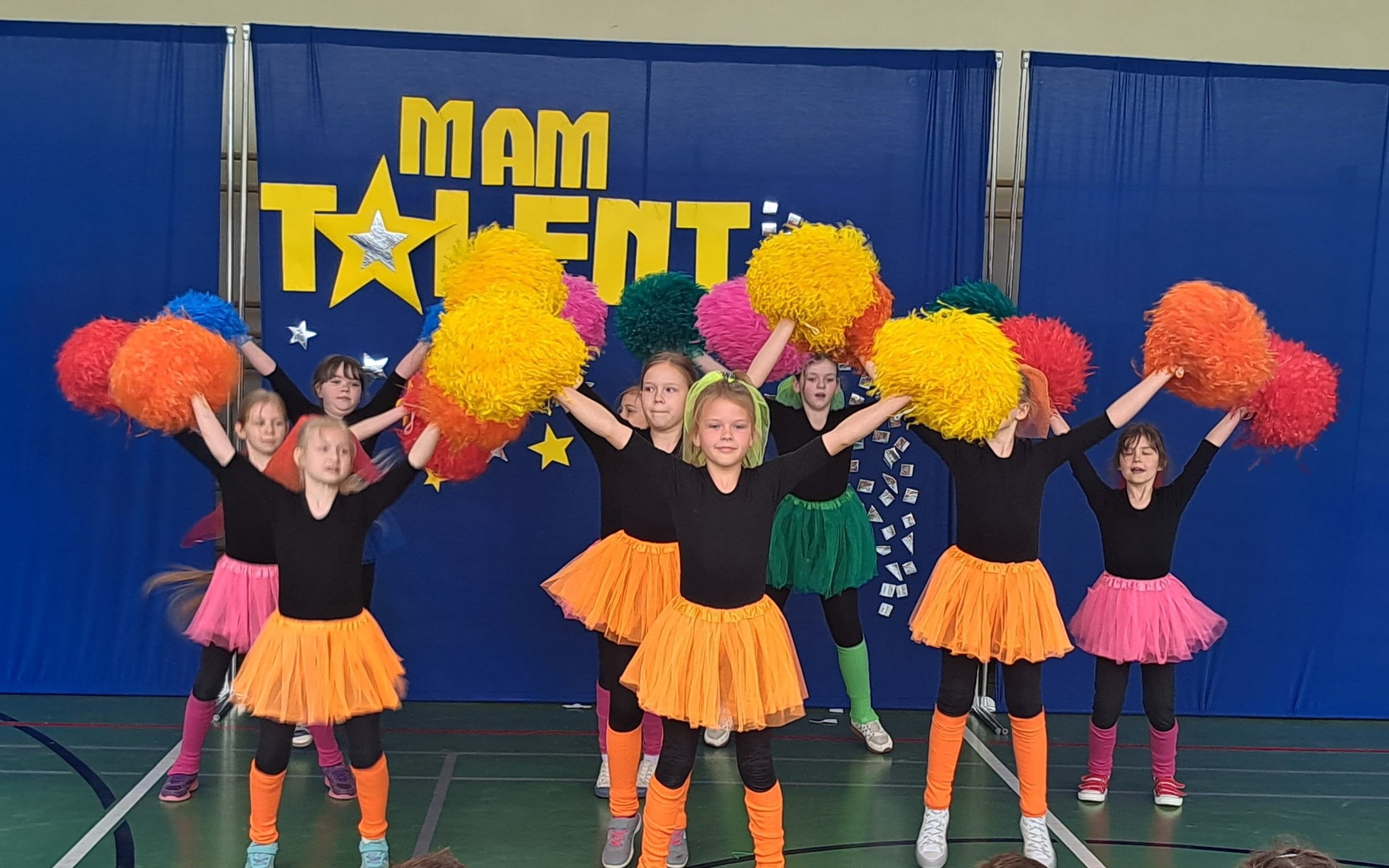 Pokaz "Mam talent" Grupa dziewczynek występuje na scenie podczas szkolnej edycji programu &bdquo;Mam Talent&rdquo;. Ubrane są w czarne koszulki, kolorowe tiulowe sp&oacute;dniczki oraz legginsy i opaski w intensywnych barwach. Każda z nich trzyma kolorowe pompony w dłoniach. W tle widać niebieski parawan z ż&oacute;łtym napisem &bdquo;MAM TALENT&rdquo; oraz dekoracjami w postaci gwiazdek.