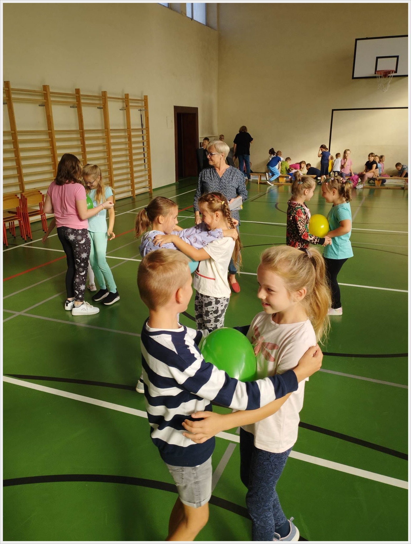 Zajęcia sportowe świetliczak&oacute;w. Dzieci biorą udział w zabawie ruchowej w sali gimnastycznej. W parach trzymają balony pomiędzy swoimi ciałami, ćwicząc wsp&oacute;łpracę i r&oacute;wnowagę. W tle widoczne są inne grupy dzieci, nauczyciele oraz sprzęt sportowy. Na ścianie znajduje się drabinka gimnastyczna i tablica do koszyk&oacute;wki. Atmosfera jest radosna i aktywna.