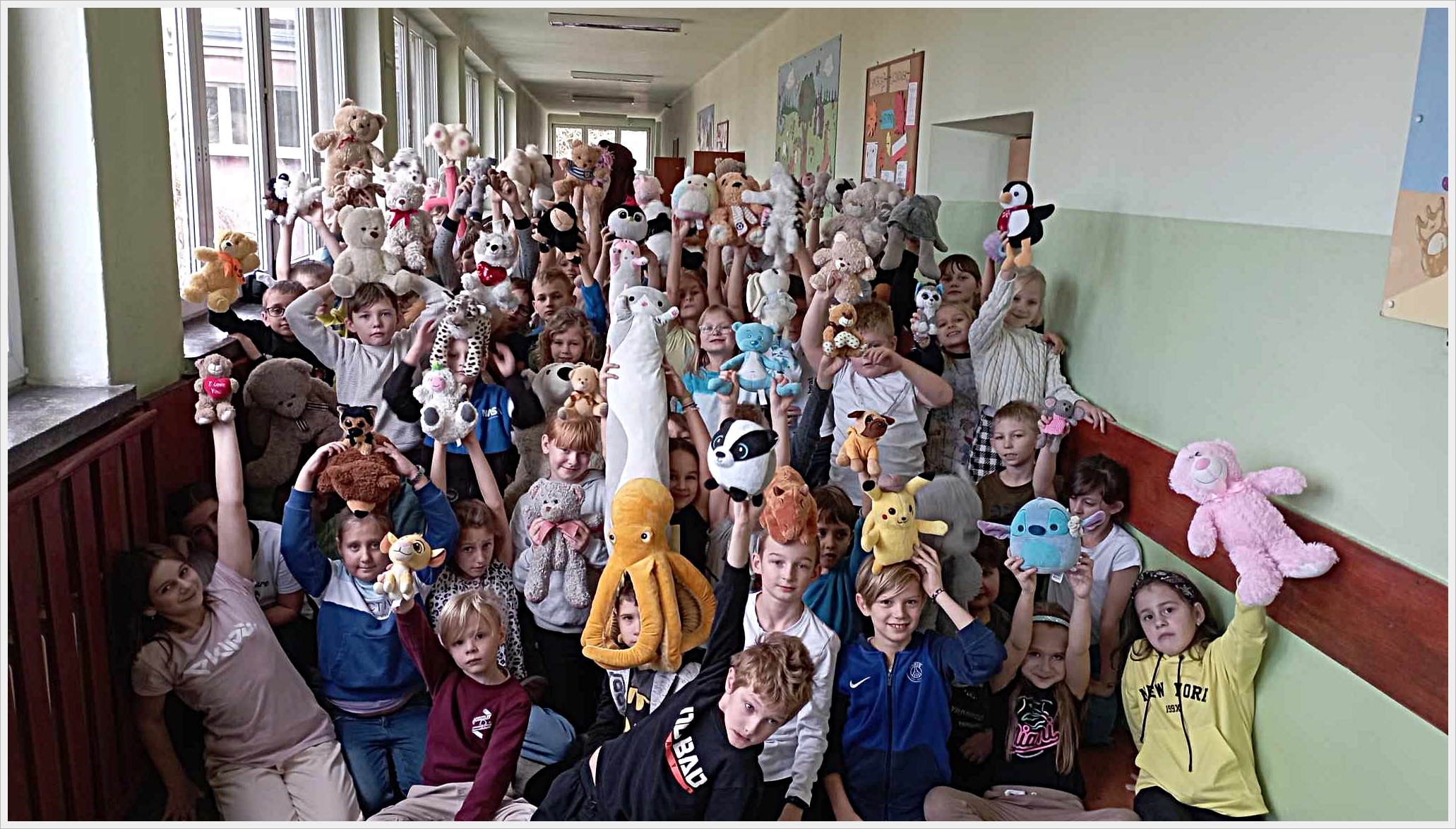 Dzień pluszaka. Grupa dzieci siedzi i stoi w szkolnym korytarzu, trzymając w g&oacute;rze swoje pluszowe maskotki. Dzieci są uśmiechnięte i radosne, a maskotki są r&oacute;żnorodne &ndash; od misi&oacute;w, przez pingwiny i koty, po ośmiornice i postacie z bajek. Scena odbywa się w jasnym korytarzu z dużymi oknami po lewej stronie. W tle widać kolorowe plakaty na ścianach.