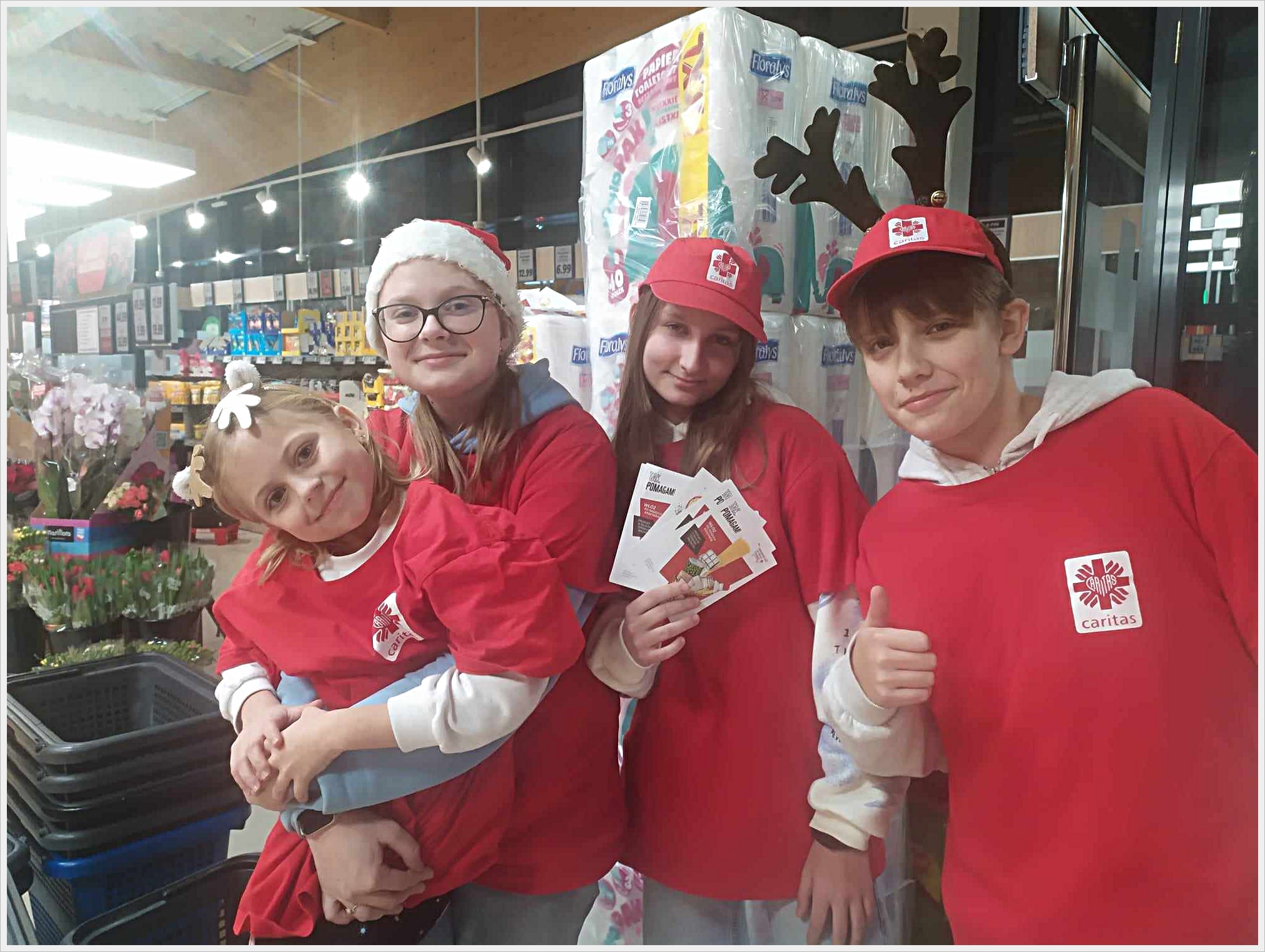 Wolontariusz naszej szkoły. Czworo dzieci ubranych w czerwone koszulki z logo Caritas pozuje do zdjęcia w sklepie spożywczym. Jedna dziewczynka trzyma na rękach młodszą dziewczynkę z opaską w kształcie kwiatka. Druga dziewczynka trzyma w rękach ulotki, a chłopiec po prawej stronie pokazuje gest &bdquo;ok&rdquo; z uniesionym kciukiem. Dzieci mają na głowach świąteczne akcenty &ndash; czapki Mikołaja i opaskę z rogami renifera. W tle widać kwiaty, w&oacute;zki sklepowe i regały z produktami.