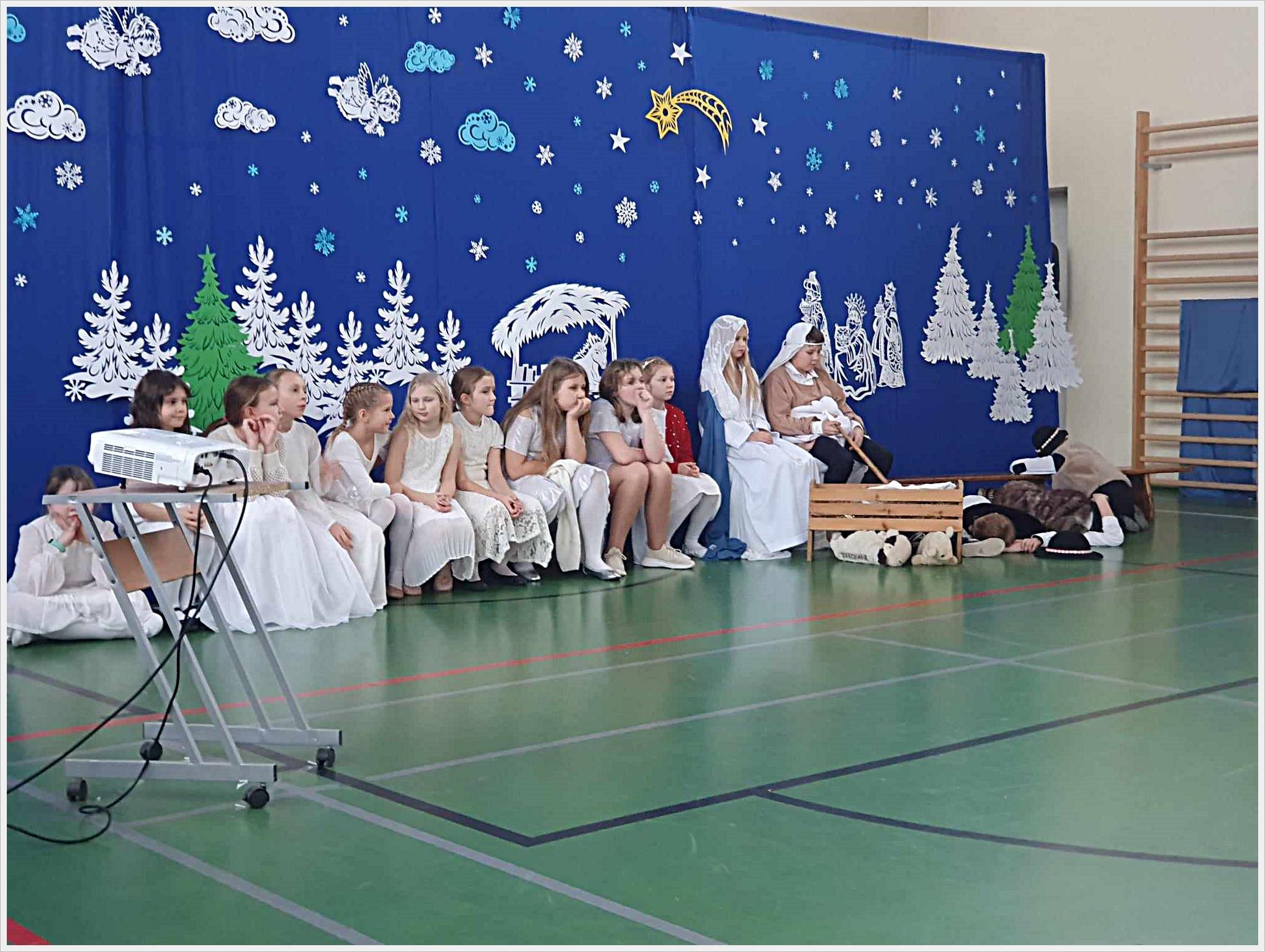 Jasełka szkolne. Grupa dzieci bierze udział w przedstawieniu jasełkowym w sali gimnastycznej. Na tle niebieskiej dekoracji z motywami zimowymi (gwiazdy, płatki śniegu, chmurki, aniołki, choinki) dzieci siedzą na ławce lub na podłodze. Po prawej stronie scena z Maryją, J&oacute;zefem i lalką w roli Dzieciątka Jezus w drewnianym żł&oacute;bku, otoczona przez dzieci przebrane za pasterzy i zwierzęta. Przed grupą stoi projektor na metalowym stoliku. Całość utrzymana w świątecznym, zimowym klimacie.