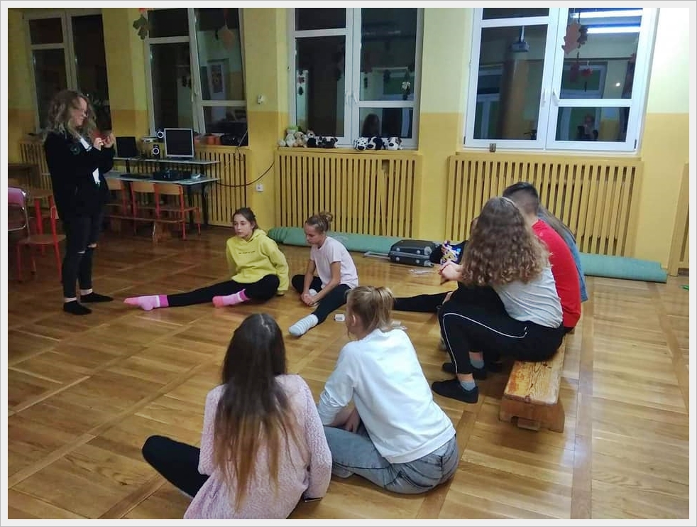 Nocowanie w szkole. Grupa młodzieży siedzi na podłodze w świetlicy lub sali szkolnej, rozmawiając i spędzając wsp&oacute;lnie czas. Część uczni&oacute;w siedzi na drewnianej ławce, a pozostali bezpośrednio na drewnianym parkiecie. W tle widoczne są biurka, komputer, głośniki oraz parapety z maskotkami. Okna są duże, z białymi ramami, a ściany pomalowane na jasnoż&oacute;łty kolor. W pomieszczeniu panuje wieczorna atmosfera, a światło pochodzi z oświetlenia wewnętrznego.