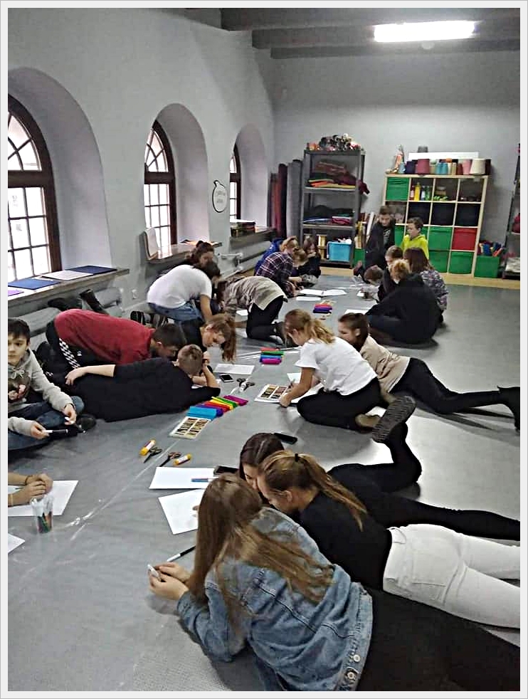 Niecodzienne warsztaty. Grupa dzieci i młodzieży leży i siedzi na podłodze w dużej sali z łukowatymi oknami, wykonując prace plastyczne. Uczniowie rysują i kolorują na kartkach, używając flamastr&oacute;w i kredek, a wok&oacute;ł nich rozłożone są przybory szkolne. W tle widać p&oacute;łki z kolorowymi pojemnikami i materiałami edukacyjnymi. Panuje tw&oacute;rcza, skupiona atmosfera.