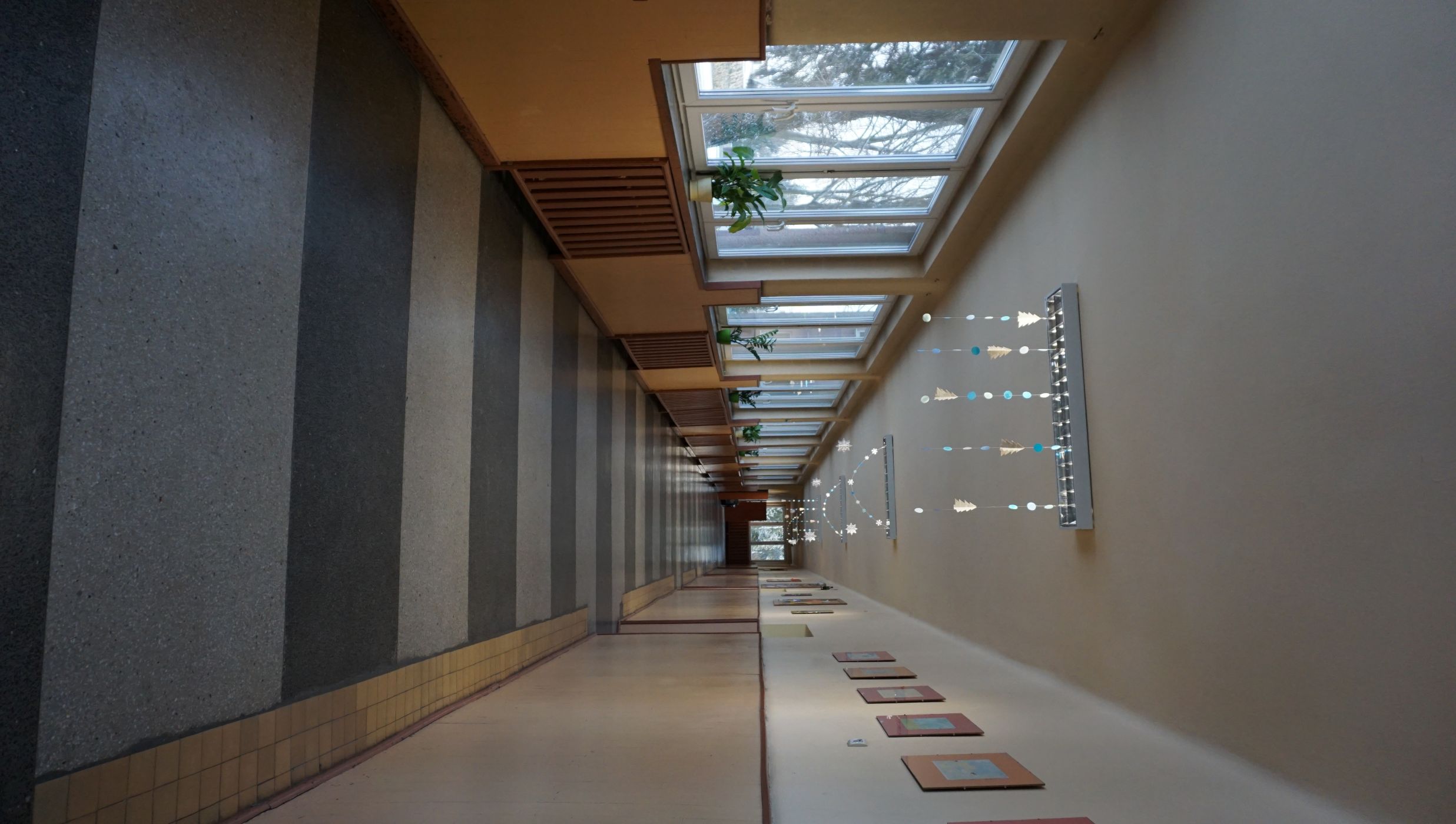 Korytarz na parterze. Długi, jasny korytarz szkolny z pasami szarej i ciemnoszarej wykładziny na podłodze. Po lewej stronie znajdują się duże okna z białymi ramami, przez kt&oacute;re widać drzewa i zewnętrzną część szkoły. Na parapetach stoją doniczki z zielonymi roślinami. Na suficie wiszą ozdoby w kształcie płatk&oacute;w śniegu i choinek, wykonane z papieru i koralik&oacute;w, zawieszone na nitkach pod lampami. Prawa ściana ozdobiona jest r&oacute;wno rozmieszczonymi kolorowymi rysunkami dzieci. Korytarz jest pusty i dobrze oświetlony naturalnym światłem dziennym.