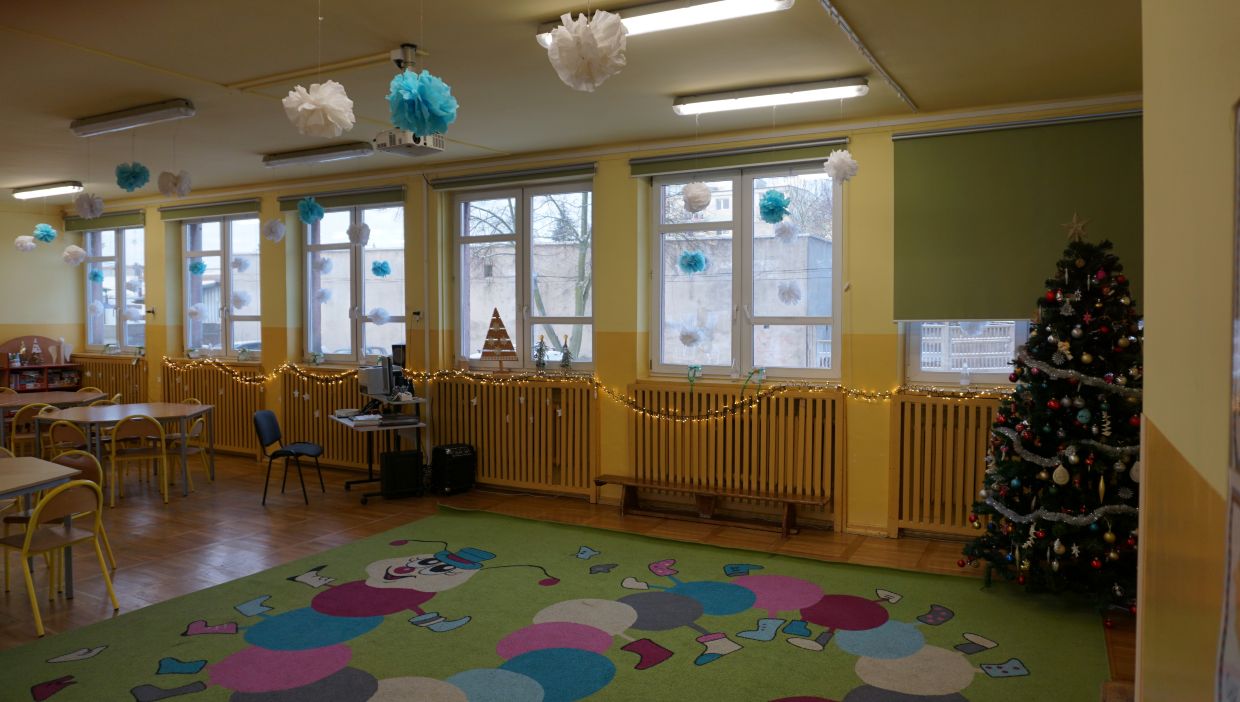 Świetlica szkoolna. Kolorowa sala udekorowana na święta. Na podłodze znajduje się dywan z rysunkiem uśmiechniętej gąsienicy i kolorowych k&oacute;łek. Wzdłuż okien rozwieszone są świąteczne lampki oraz papierowe ozdoby w kolorach białym i niebieskim. W rogu sali stoi przystrojona choinka z bombkami i łańcuchami. Przy oknach znajdują się biurka i krzesła oraz komputer. Pomieszczenie ma jasnoż&oacute;łte ściany i duże okna.