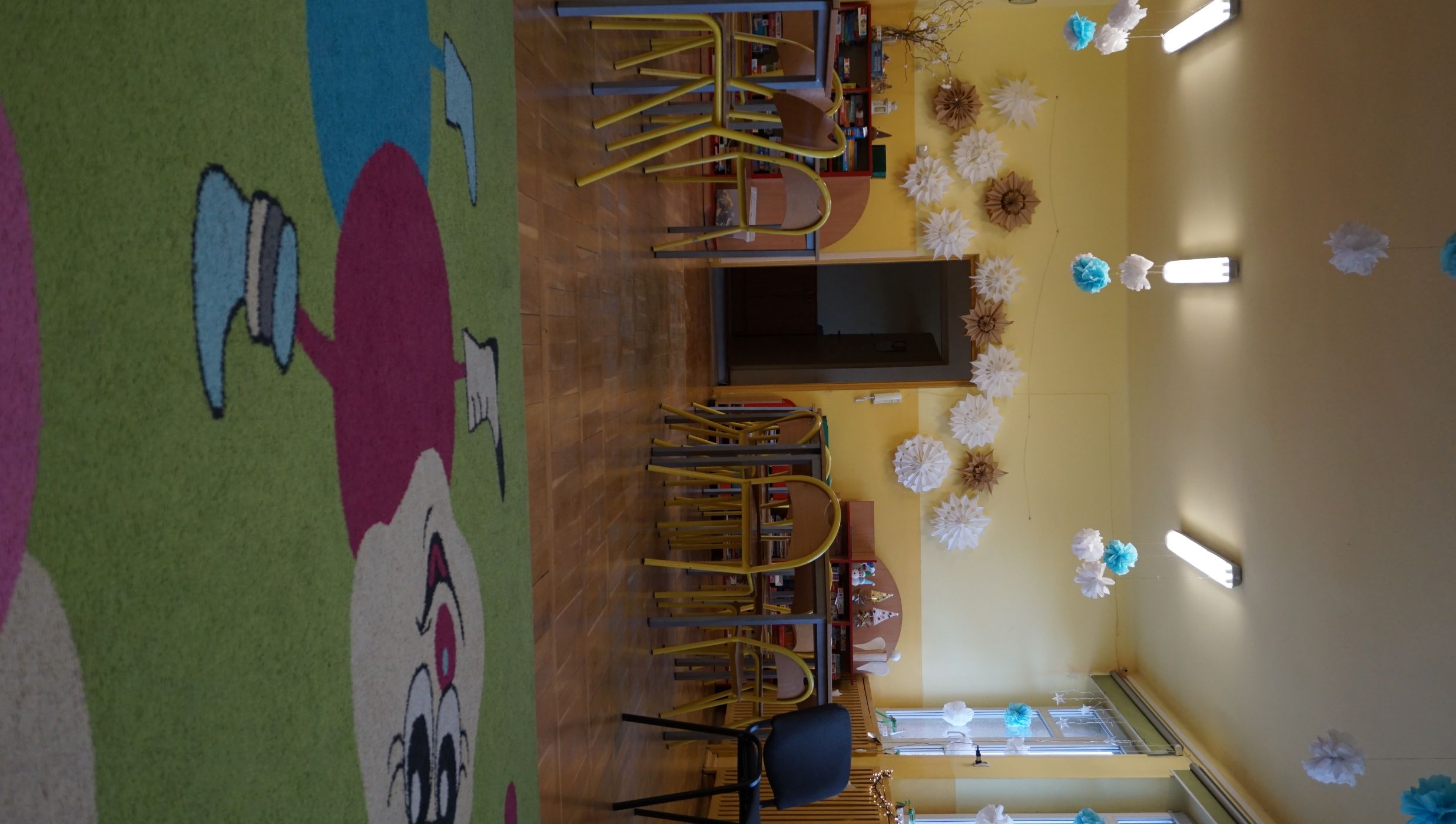 Świetlica szkolna. Sala z jasnoż&oacute;łtymi ścianami i drewnianą podłogą. Na pierwszym planie widoczny jest kolorowy dywan z rysunkiem uśmiechniętej postaci z dużymi okrągłymi segmentami ciała. W tle znajdują się dziecięce stoliki i krzesła oraz regały z grami i zabawkami. Na ścianie nad drzwiami zawieszone są dekoracje przypominające papierowe śnieżynki w kolorach białym i brązowym. Z sufitu zwisają białe i niebieskie pompony. Po prawej stronie &ndash; okna z ozdobami i lampkami.