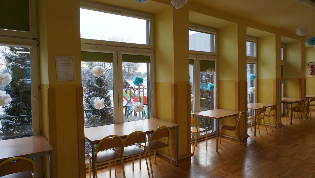 Świetlica szkolna. Jasna, ż&oacute;łta sala z dużymi oknami i drzwiami balkonowymi wychodzącymi na plac zabaw z kolorowymi zjeżdżalniami, częściowo zasłoniętymi śnieżnymi drzewami. Przy każdym oknie ustawione są proste drewniane stoliki z krzesłami. Na szybach zawieszone są papierowe dekoracje w kształcie białych i niebieskich pompon&oacute;w, nadające wnętrzu zimowy klimat. Na ścianie widoczny jest certyfikat lub dyplom, a w tle &ndash; gaśnica. Podłoga z drewnianych paneli, całość sprawia wrażenie stoł&oacute;wki lub świetlicy w plac&oacute;wce edukacyjnej.