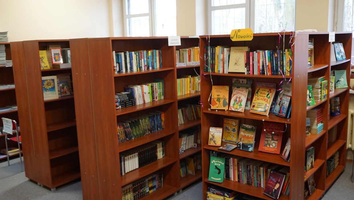 Biblioteka szkolna. Wnętrze szkolnej biblioteki z kilkoma drewnianymi regałami wypełnionymi kolorowymi książkami. Na jednym z regał&oacute;w znajduje się ż&oacute;łty szyld z napisem &bdquo;Nowości&rdquo; oraz ozdobne serpentyny. Książki są ułożone pionowo i frontem do przodu &ndash; wśr&oacute;d nich widoczne są bajki, encyklopedie, książki przygodowe i edukacyjne. W tle jasne ściany i duże okna wpuszczające naturalne światło.