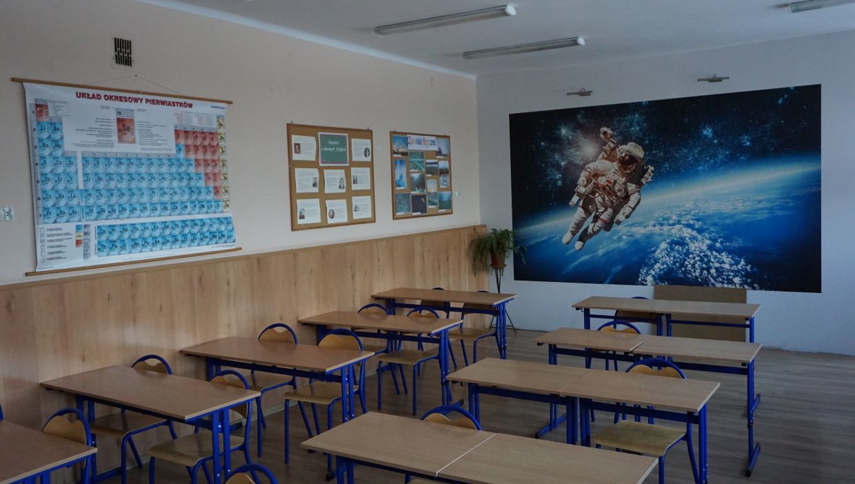 Sala fizyczna. Sala lekcyjna z ustawionymi w rzędach ławkami i krzesłami. Na ścianie po lewej stronie wisi kolorowy układ okresowy pierwiastk&oacute;w oraz tablice informacyjne. Na ścianie z tyłu znajduje się duża fototapeta przedstawiająca astronautę w przestrzeni kosmicznej nad Ziemią.