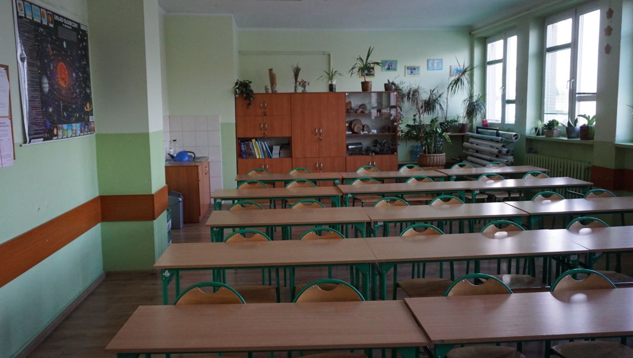 Sala geograficzna. Sala lekcyjna z rzędami pustych ławek i krzeseł. Po lewej stronie widoczny plakat przedstawiający Układ Słoneczny. Z tyłu klasy znajduje się szafka z książkami, eksponatami i licznymi roślinami doniczkowymi. Pod oknami po prawej stronie r&oacute;wnież ustawiono wiele roślin.