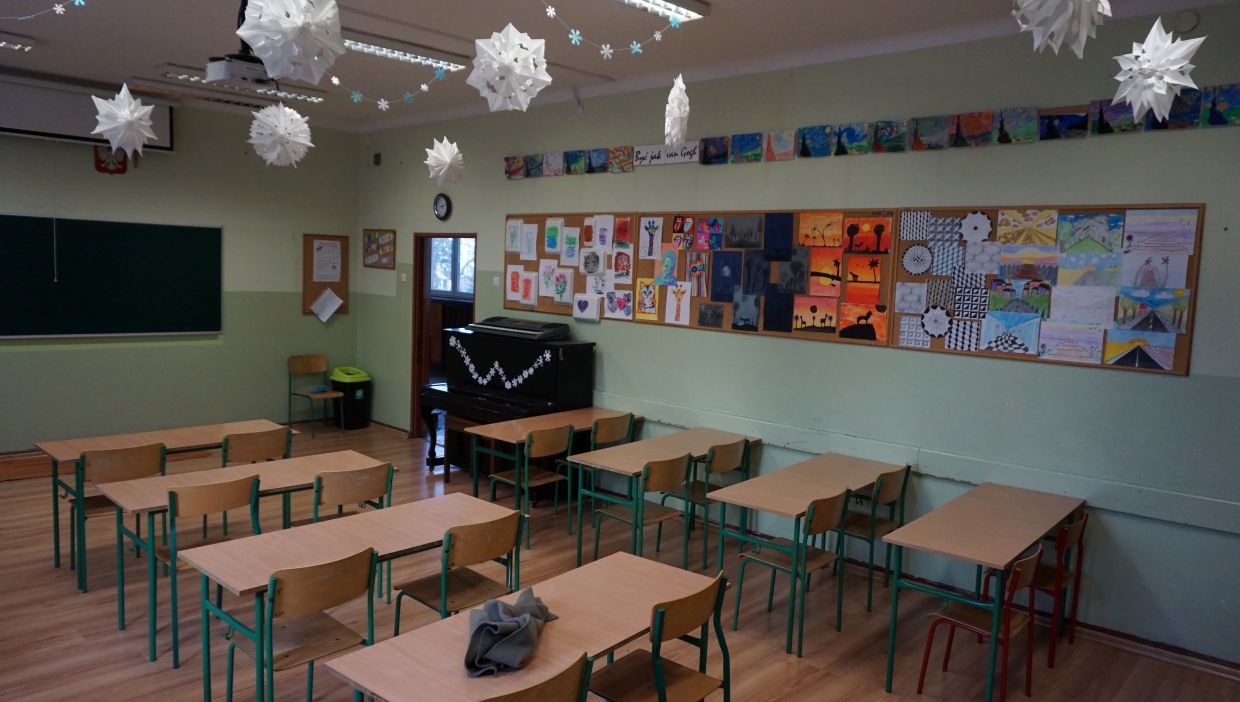 Sala języka polskiego. Sala lekcyjna z pustymi ławkami i zielonymi krzesłami. Na ścianie z prawej strony znajdują się tablice korkowe z kolorowymi rysunkami uczni&oacute;w. Nad tablicami wiszą dodatkowe prace plastyczne. Z sufitu zwisają papierowe ozdoby w kształcie śnieżynek. W rogu klasy stoi czarny pianino ozdobione śnieżynkami.